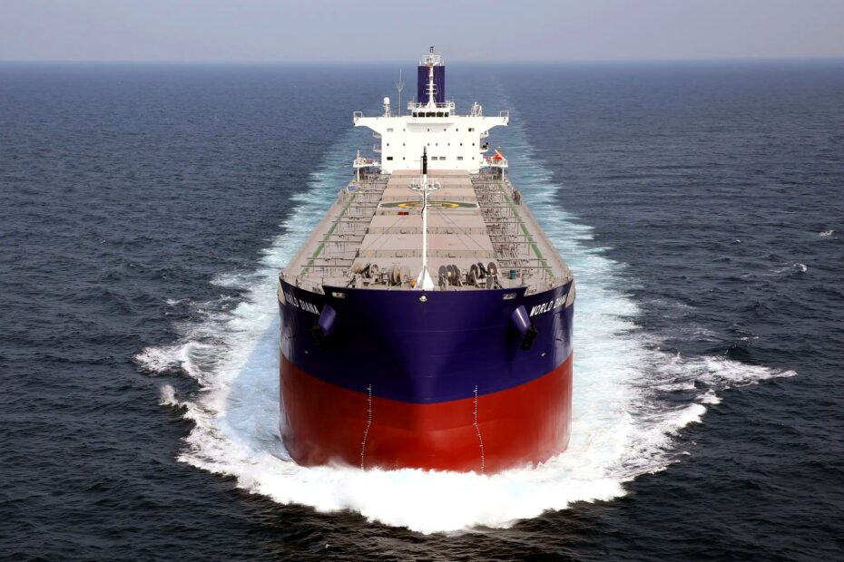 Front-facing bulk carrier sailing through open sea, leaving symmetrical white wakes beneath an overcast sky