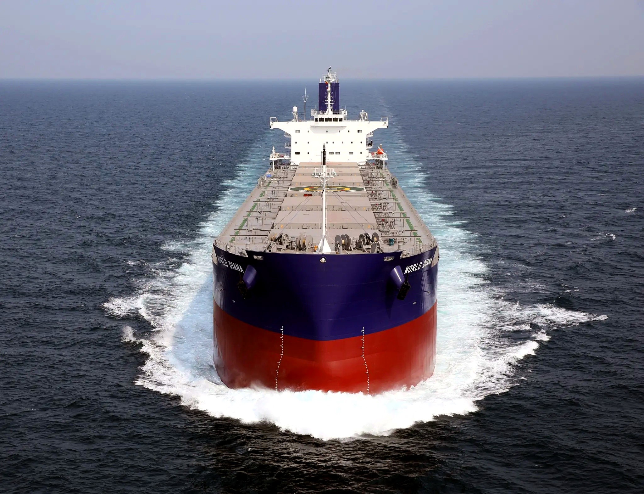 Front-facing bulk carrier sailing through open sea, leaving symmetrical white wakes beneath an overcast sky