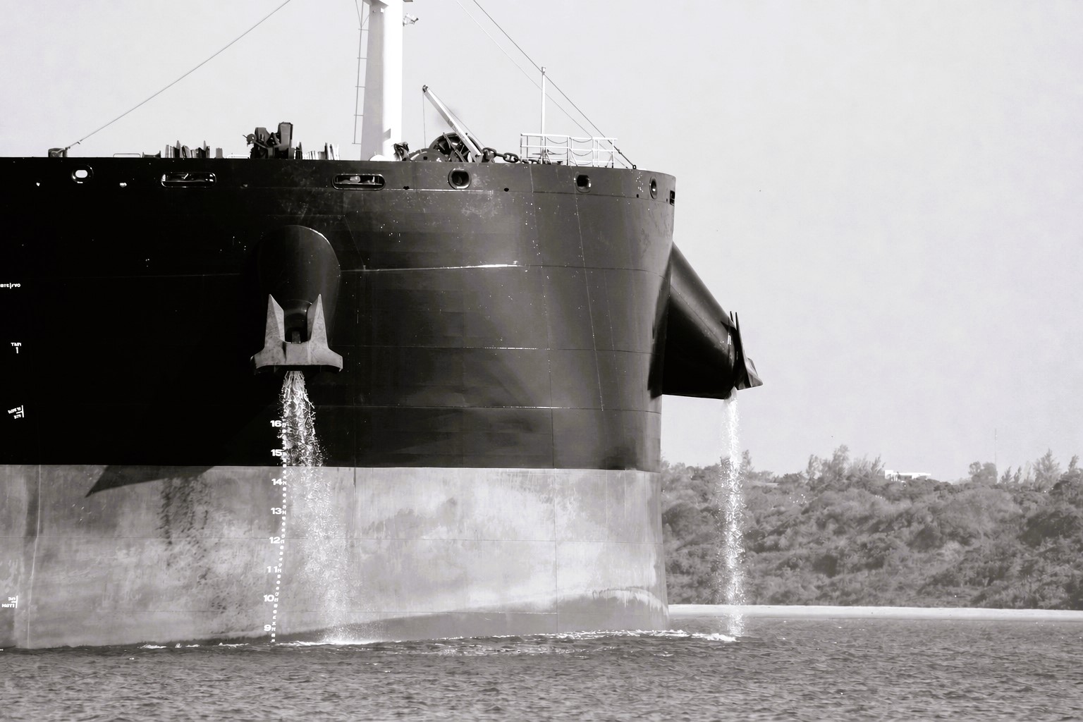 Black-and-white close-up of a large dry bulk vessel’s bow moving through calm waters, with ballast water discharging from both sides