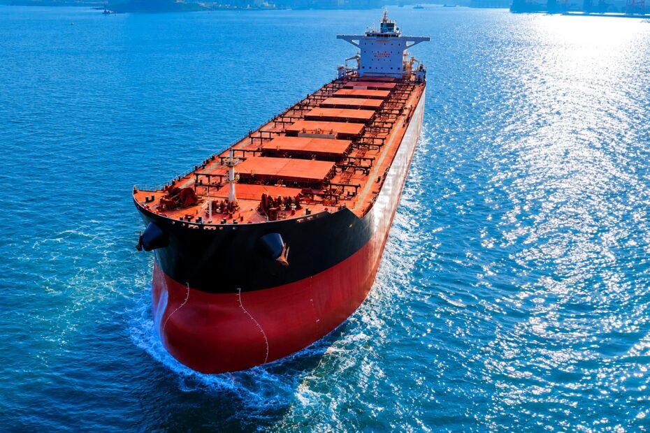 Large dry bulk carrier sailing through sunlit waters near port infrastructure, viewed from above, with vivid blue sea reflections