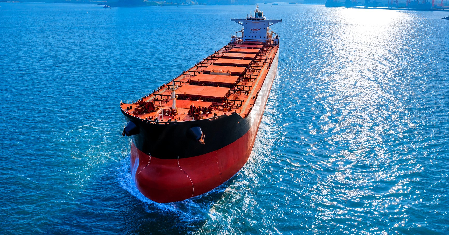 Large dry bulk carrier sailing through sunlit waters near port infrastructure, viewed from above, with vivid blue sea reflections