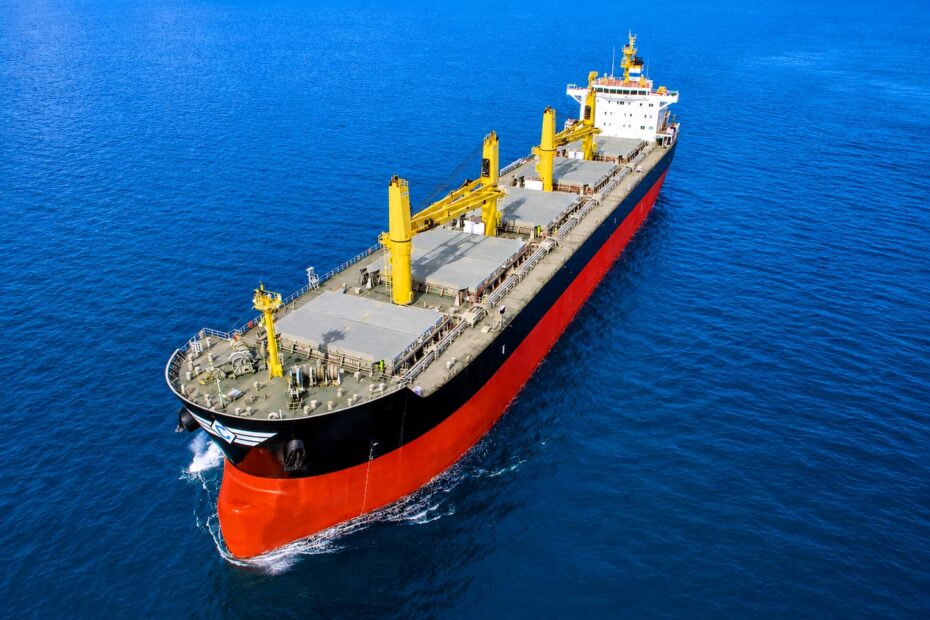 A geared dry bulk vessel sailing through deep blue waters under clear skies, viewed from above at sea