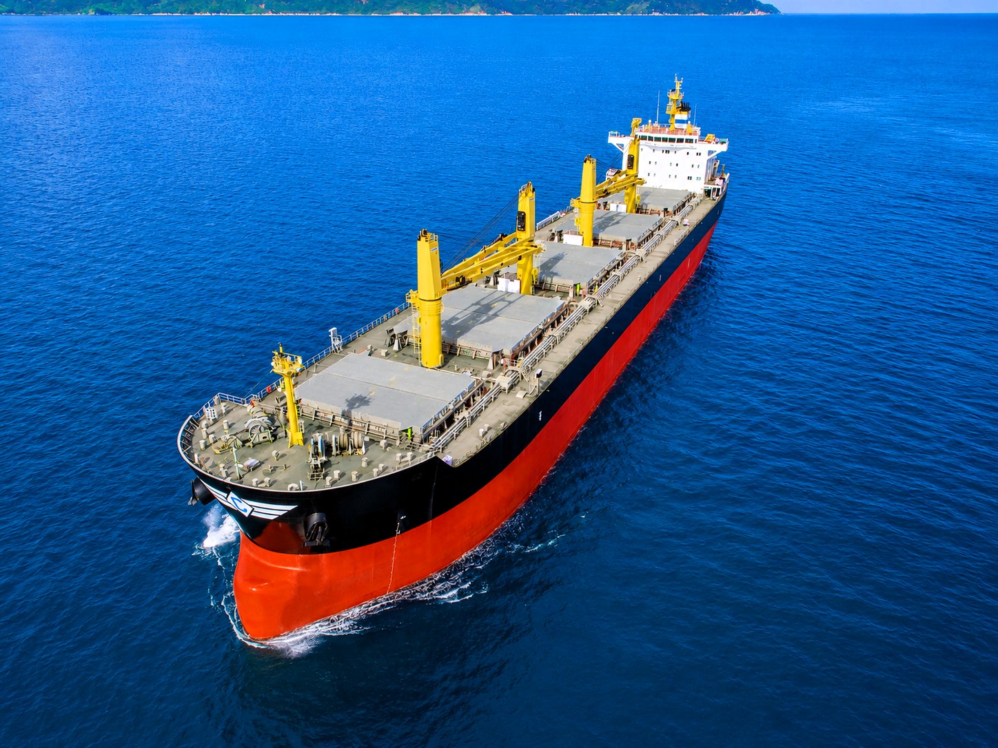 A geared dry bulk vessel sailing through deep blue waters under clear skies, viewed from above at sea