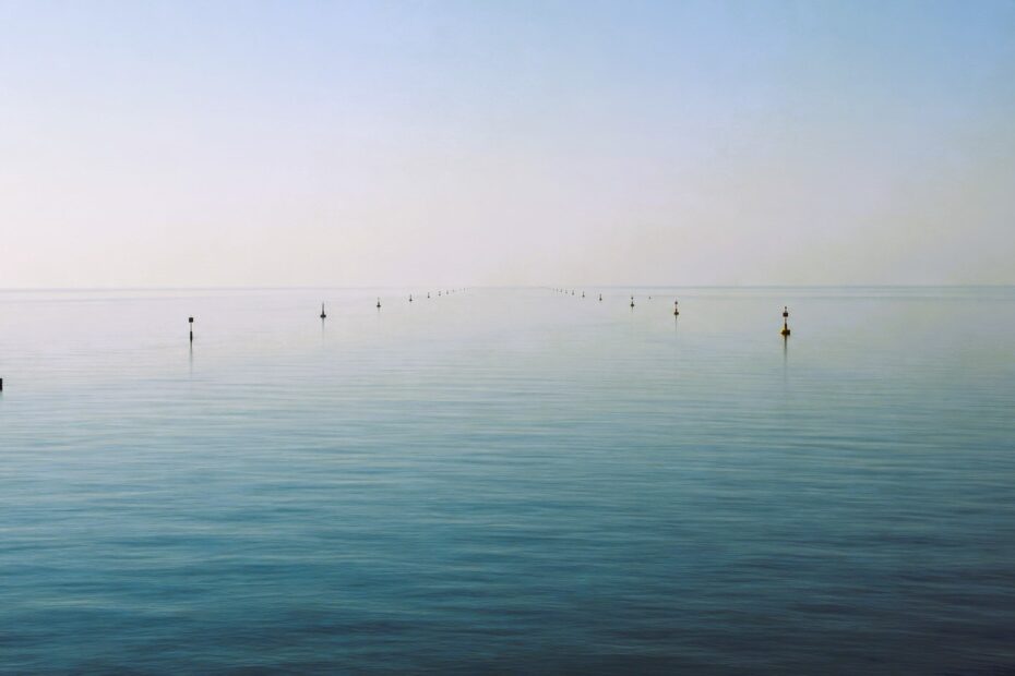 Minimalist maritime scene showing calm open water with aligned navigation buoys under hazy daylight and no vessels in transit
