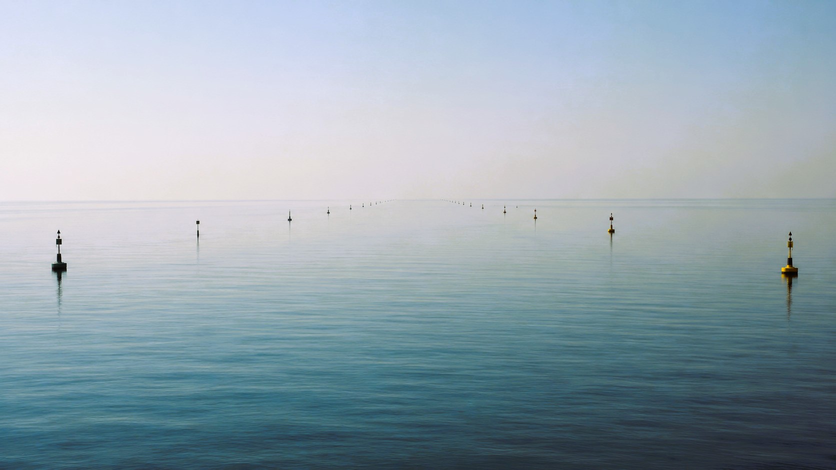 Minimalist maritime scene showing calm open water with aligned navigation buoys under hazy daylight and no vessels in transit