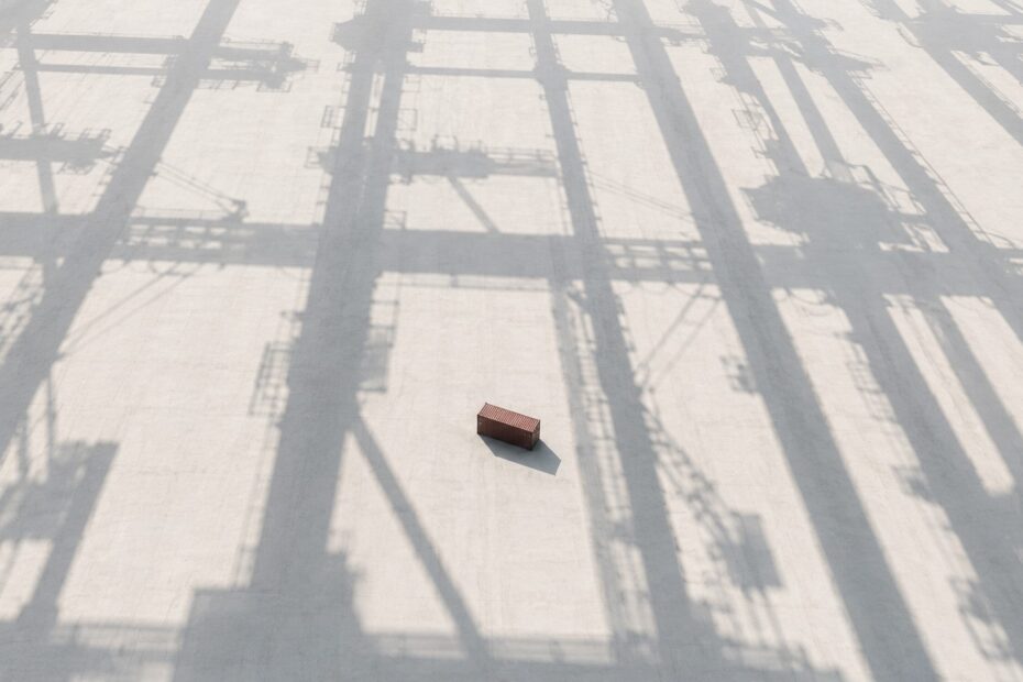 Minimalist aerial industrial landscape with geometric infrastructure shadows surrounding an isolated container, symbolising structural imbalance in global shipping networks