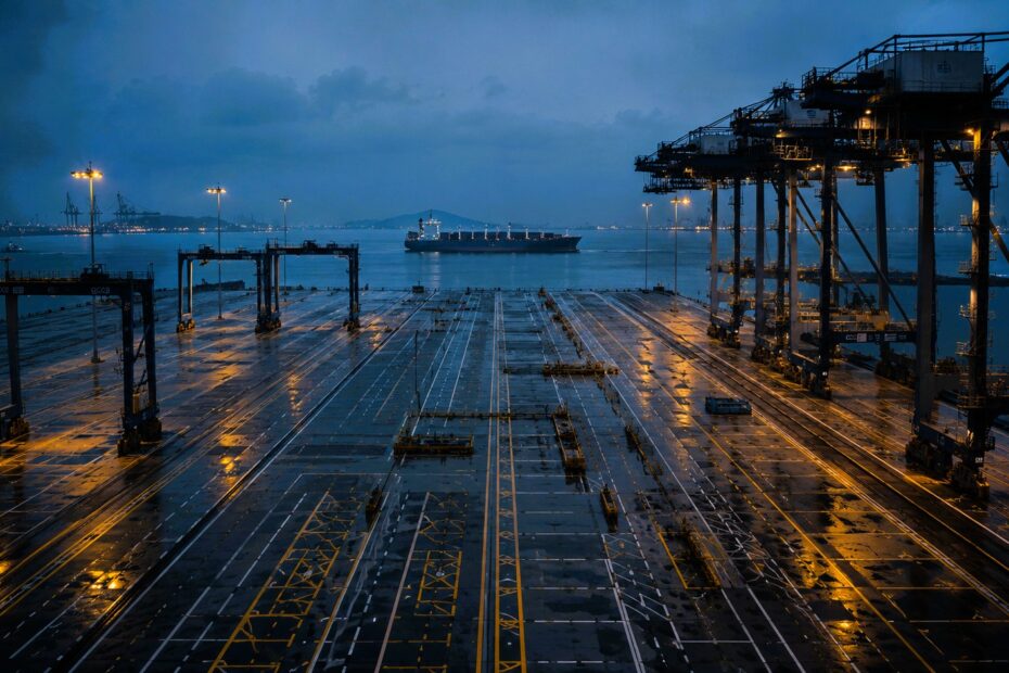 Empty automated container terminal at dusk with towering cranes, reflective concrete lanes and a distant cargo vessel on the horizon