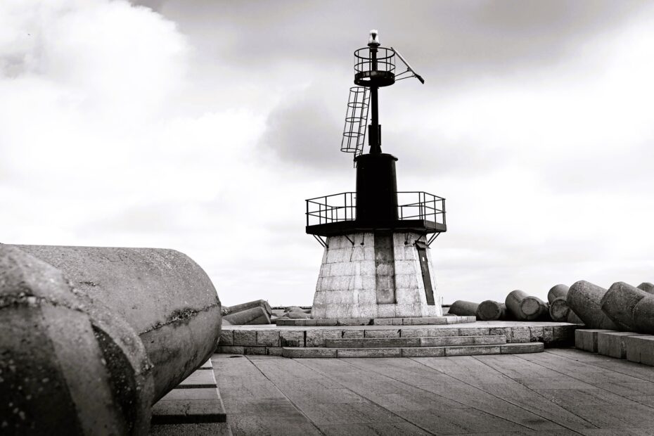 Black-and-white photograph of a lighthouse structure on a concrete breakwater, surrounded by wave barriers beneath a cloudy sky
