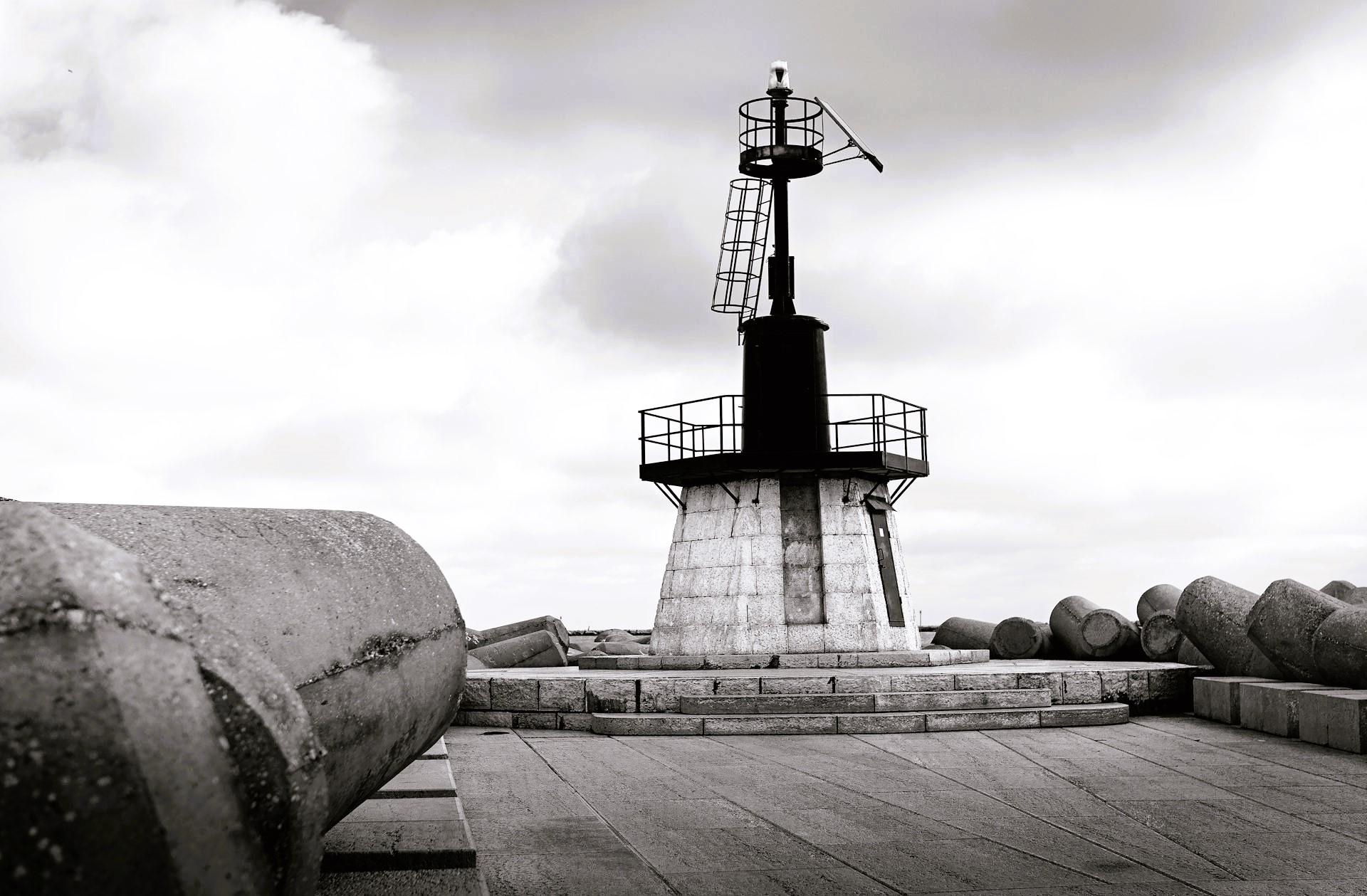 Black-and-white photograph of a lighthouse structure on a concrete breakwater, surrounded by wave barriers beneath a cloudy sky
