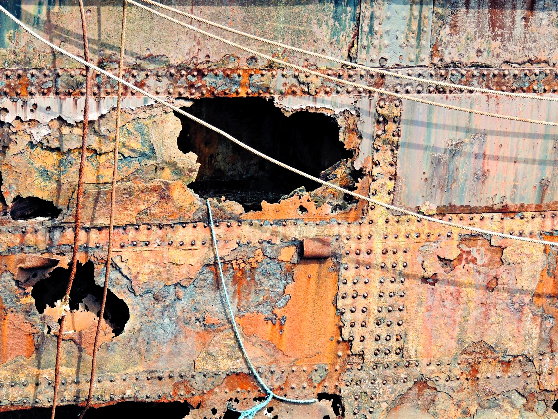 Close-up of a heavily weathered steel ship hull with extensive corrosion, peeling surfaces, riveted plates and jagged openings