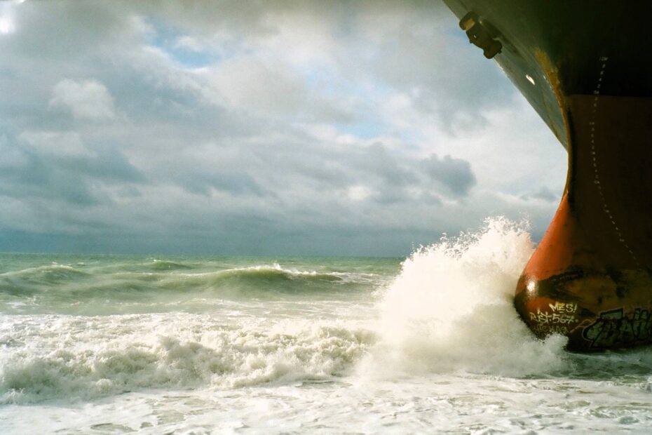 Low-angle view of a large commercial vessel’s bow cutting through rough seas beneath heavy clouds, with waves crashing against the hull