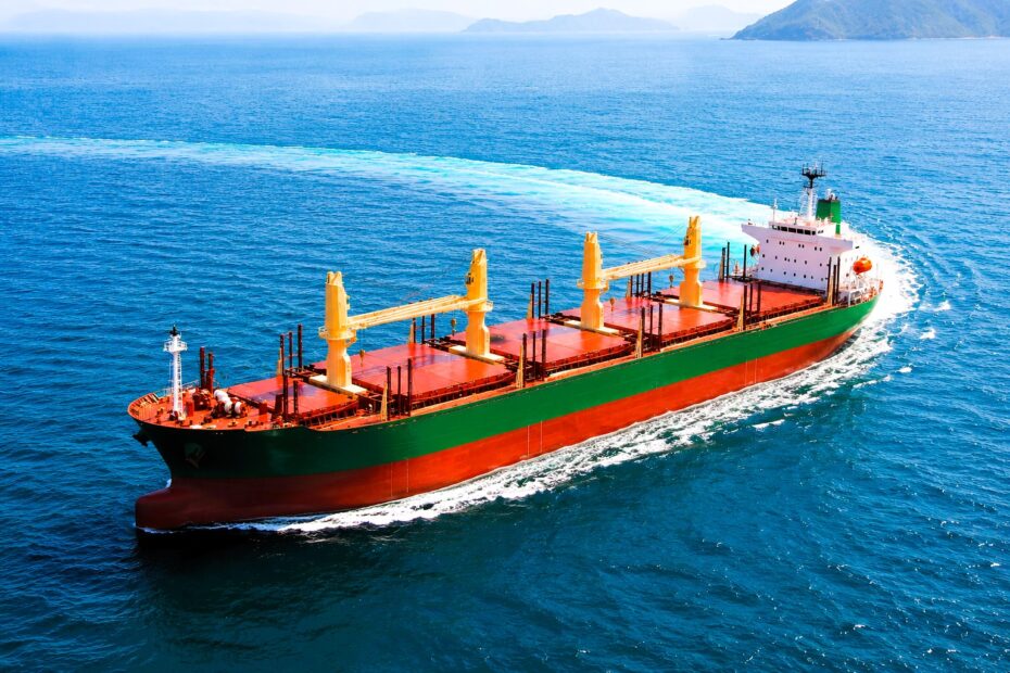A bulk carrier sails through open water under clear daylight, with enhanced colours, a green hull, and a broad wake curving across the sea