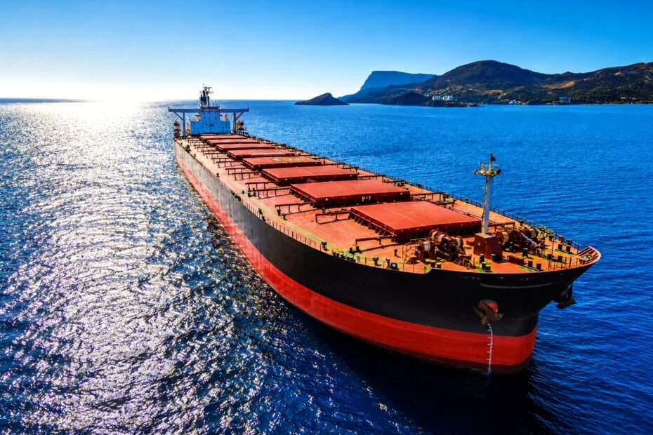 A dry bulk carrier sails across calm blue waters under bright sunlight, with distant mountainous coastline visible in the background