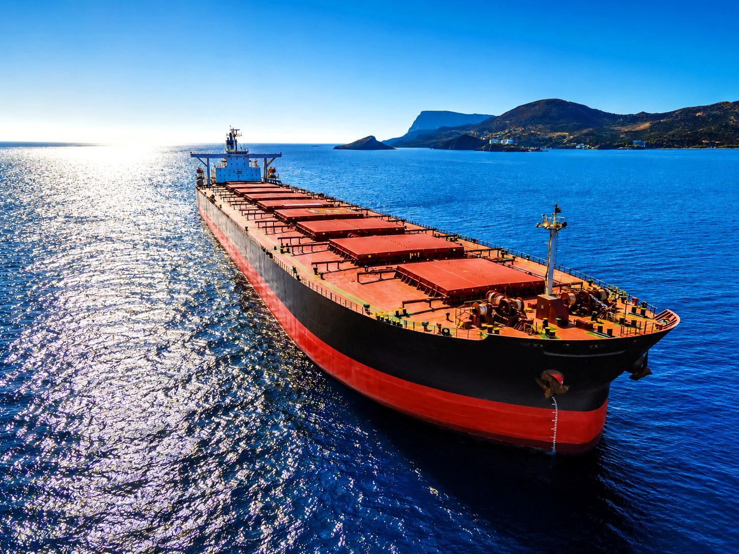 A dry bulk carrier sails across calm blue waters under bright sunlight, with distant mountainous coastline visible in the background