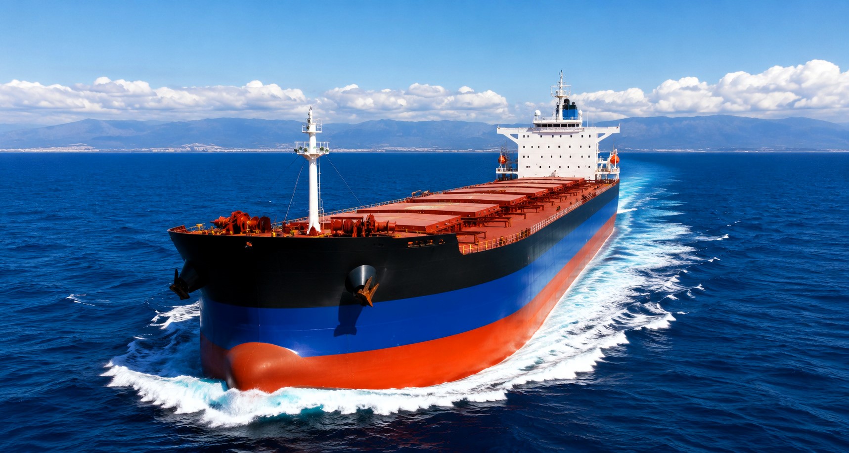 High-resolution aerial view of a bulk carrier sailing through deep blue waters under bright skies, with distant mountains on the horizon and a long white wake trailing behind the vessel
