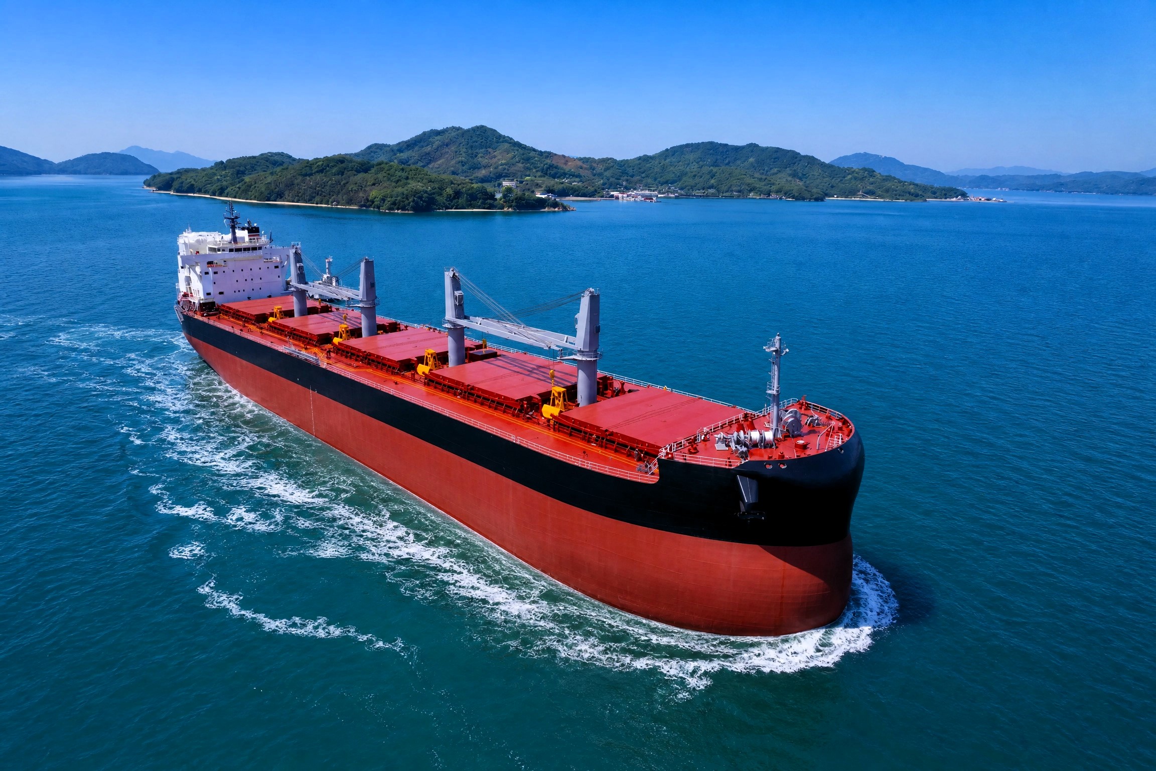 A large dry bulk vessel underway in calm blue waters, viewed from above, with green islands and distant hills in the background under clear skies