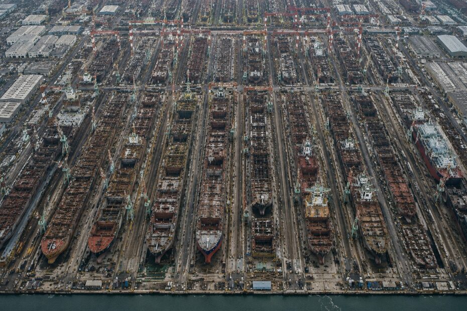 Aerial view of a vast shipyard with numerous vessels under construction in parallel dry docks, creating a dense pattern of industrial activity, cranes, infrastructure and geometric repetition stretching across the frame