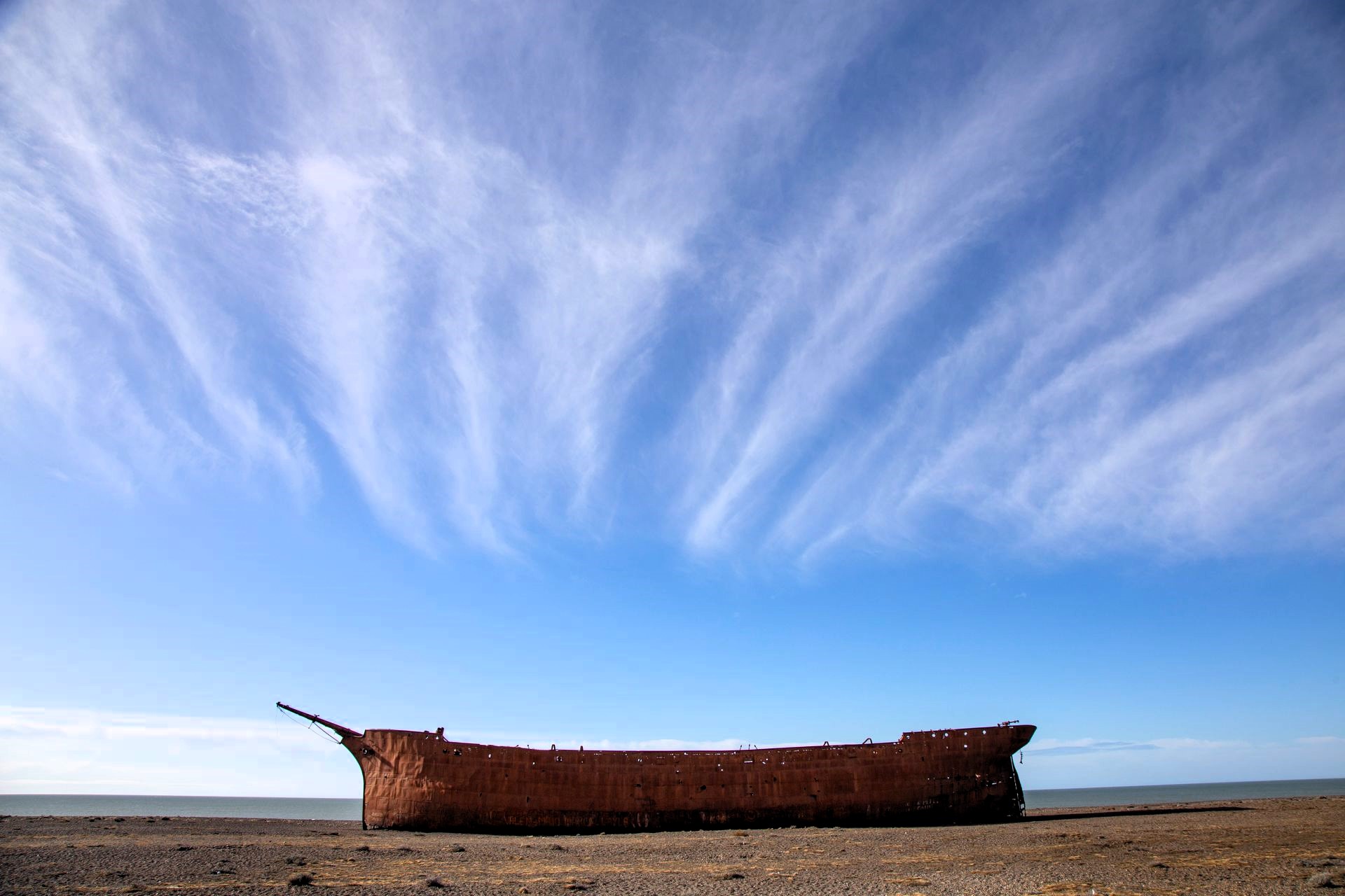 A weathered ship hull rests alone on a barren shoreline beneath sweeping high-altitude clouds stretching across a vast blue sky