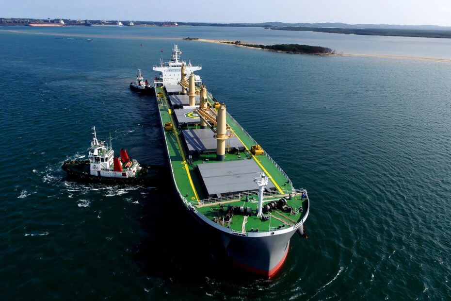 Aerial view of an Ultramax bulk carrier assisted by tugboats while navigating a coastal channel under clear skies, with calm waters and shoreline visible in the background