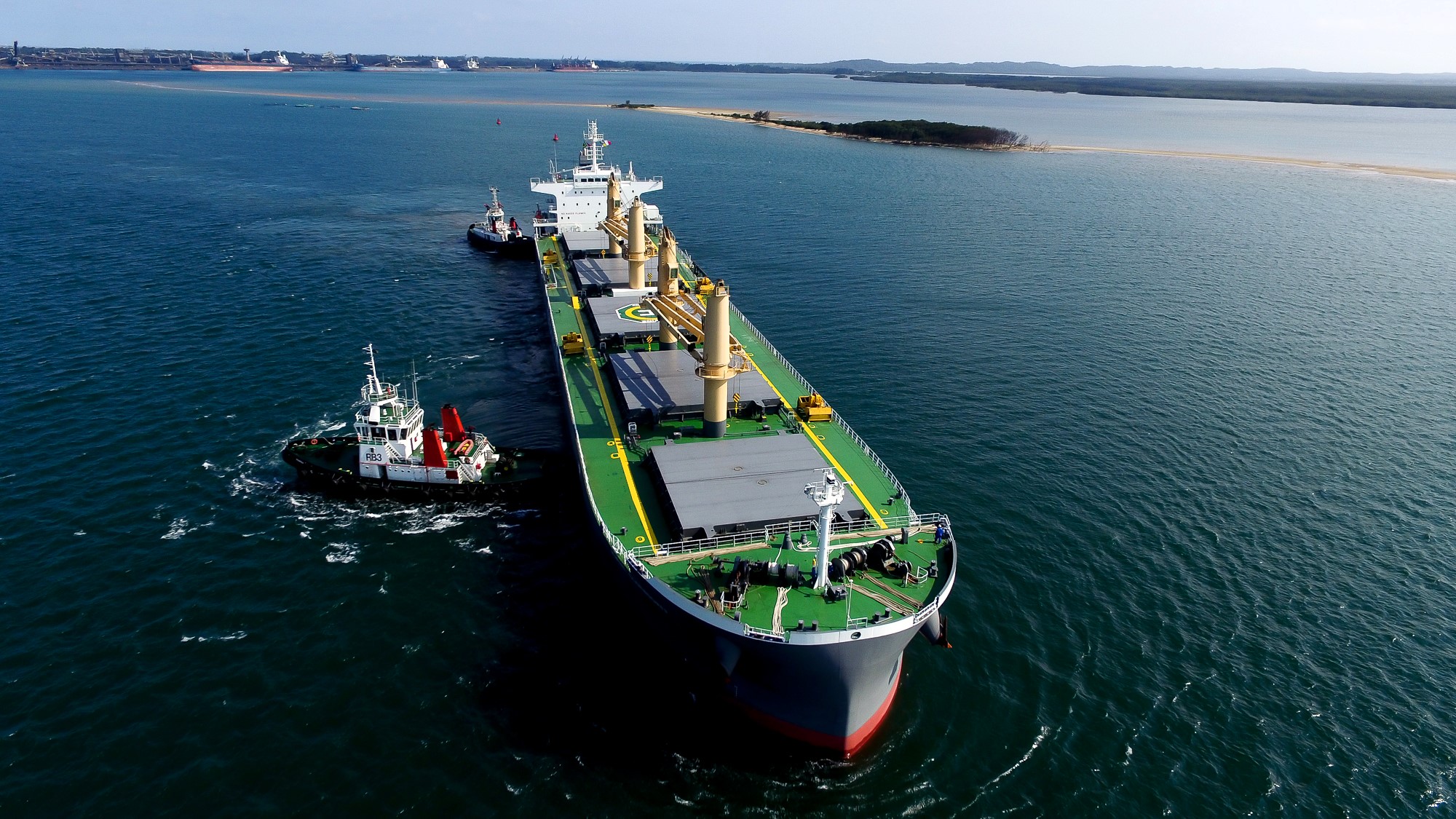 Aerial view of an Ultramax bulk carrier assisted by tugboats while navigating a coastal channel under clear skies, with calm waters and shoreline visible in the background
