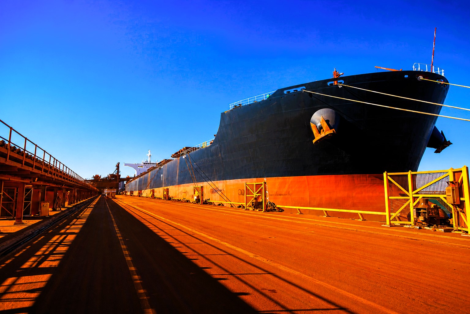 A Capesize bulk carrier berthed at an iron ore export terminal, highlighting the scale of dry bulk logistics, loading infrastructure, and global commodity flows