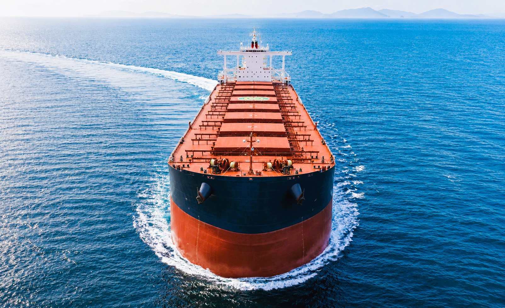 A large Capesize bulk carrier sails through calm open waters under natural daylight, leaving a broad wake with a clear horizon ahead