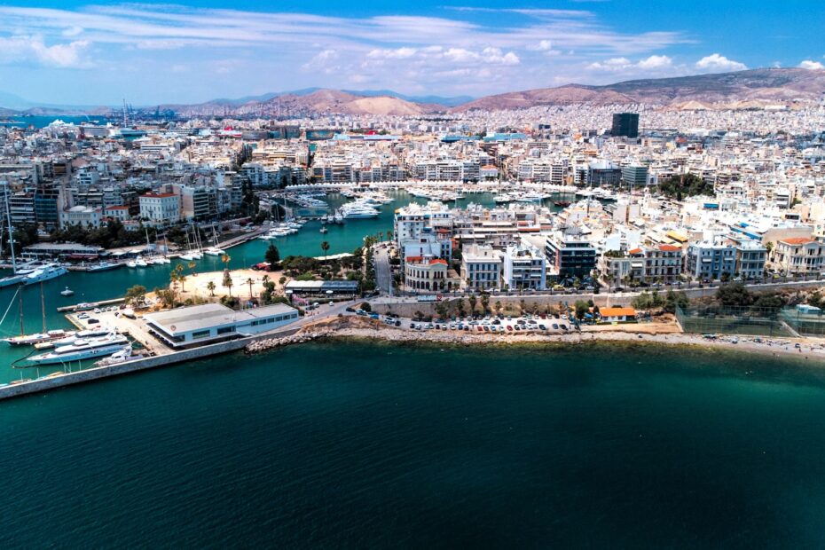 Panoramic aerial view of Piraeus, Greece, showing the marina, dense urban waterfront, commercial port facilities, and the surrounding coastline beneath a clear summer sky