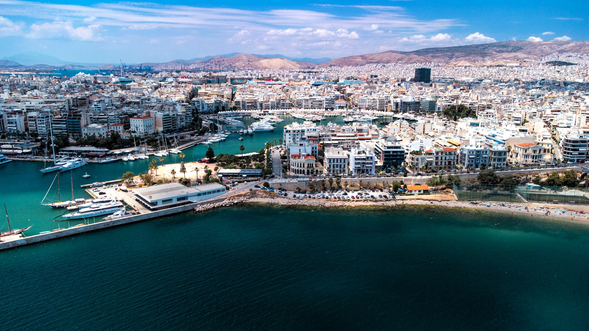 Panoramic aerial view of Piraeus, Greece, showing the marina, dense urban waterfront, commercial port facilities, and the surrounding coastline beneath a clear summer sky