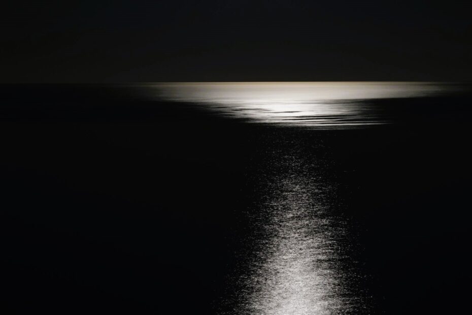 Moonlight reflecting across a calm, dark ocean at night, illuminating a silent horizon beneath an empty sky