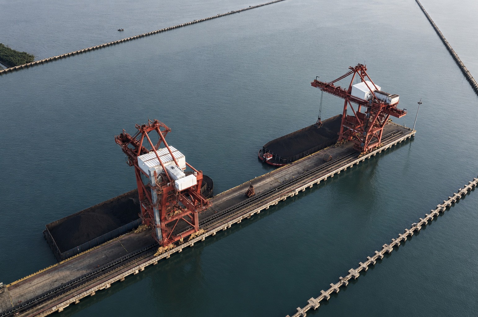 Aerial view of a dry bulk coal terminal with two large shiploaders, conveyor infrastructure, and coal-filled vessels alongside the pier