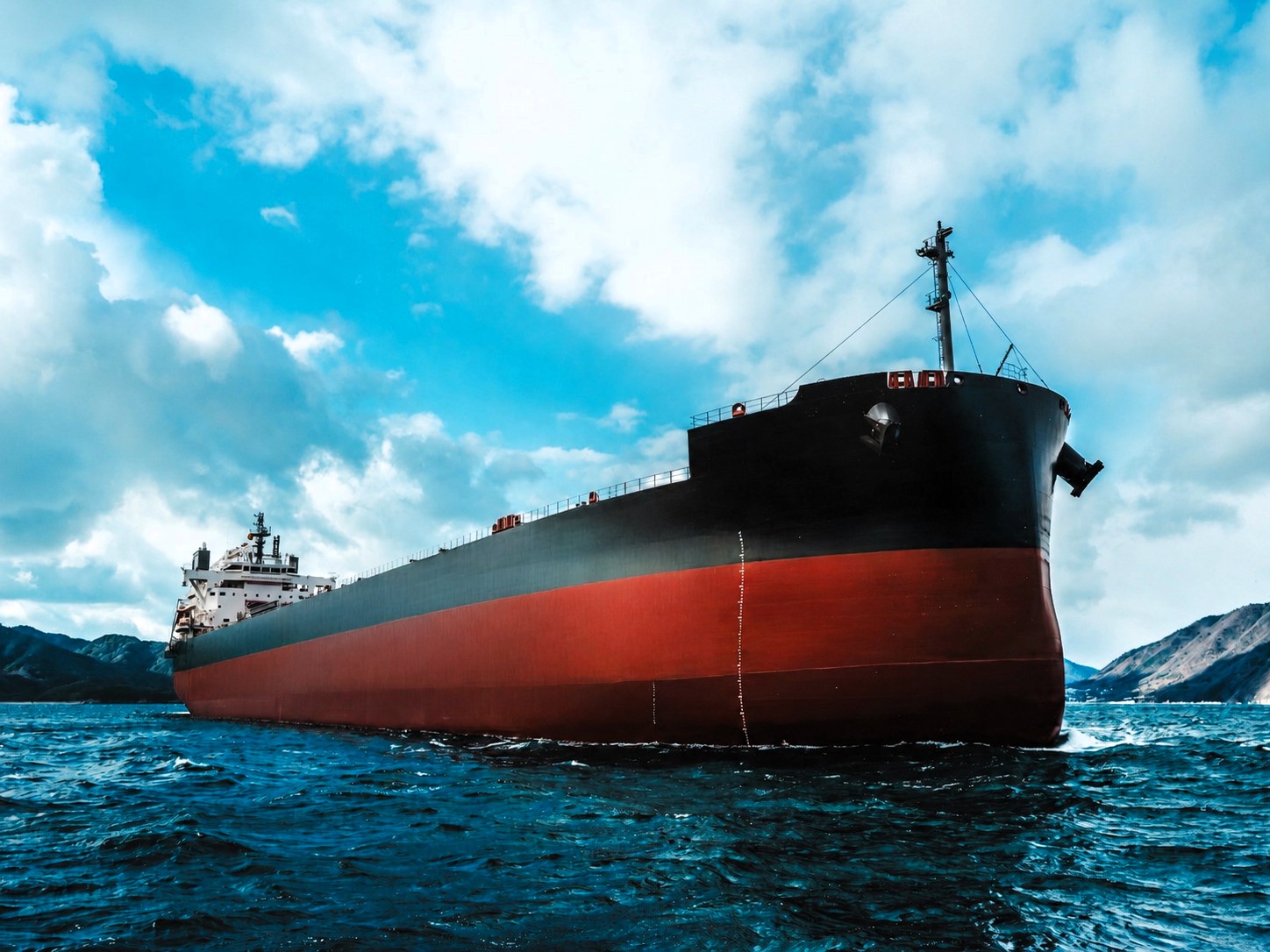 Low-angle view of a large dry bulk carrier sailing in coastal waters beneath a dramatic cloud-filled sky, with mountainous shoreline visible in the background