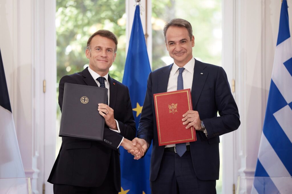 Mitsotakis and Macron shaking hands with signed agreement folders in Athens, the Hellenic bloc's extra-NATO guarantee