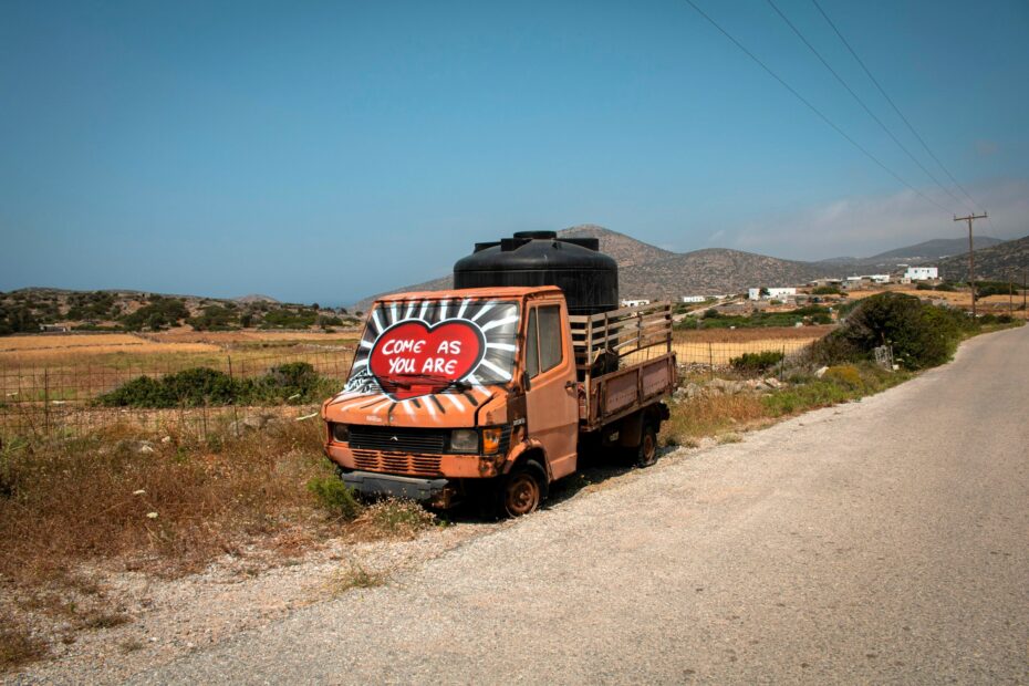 Weathered orange truck marked “Come As You Are” beside a rural road in the dry, mountainous landscape of Amorgos, Greece