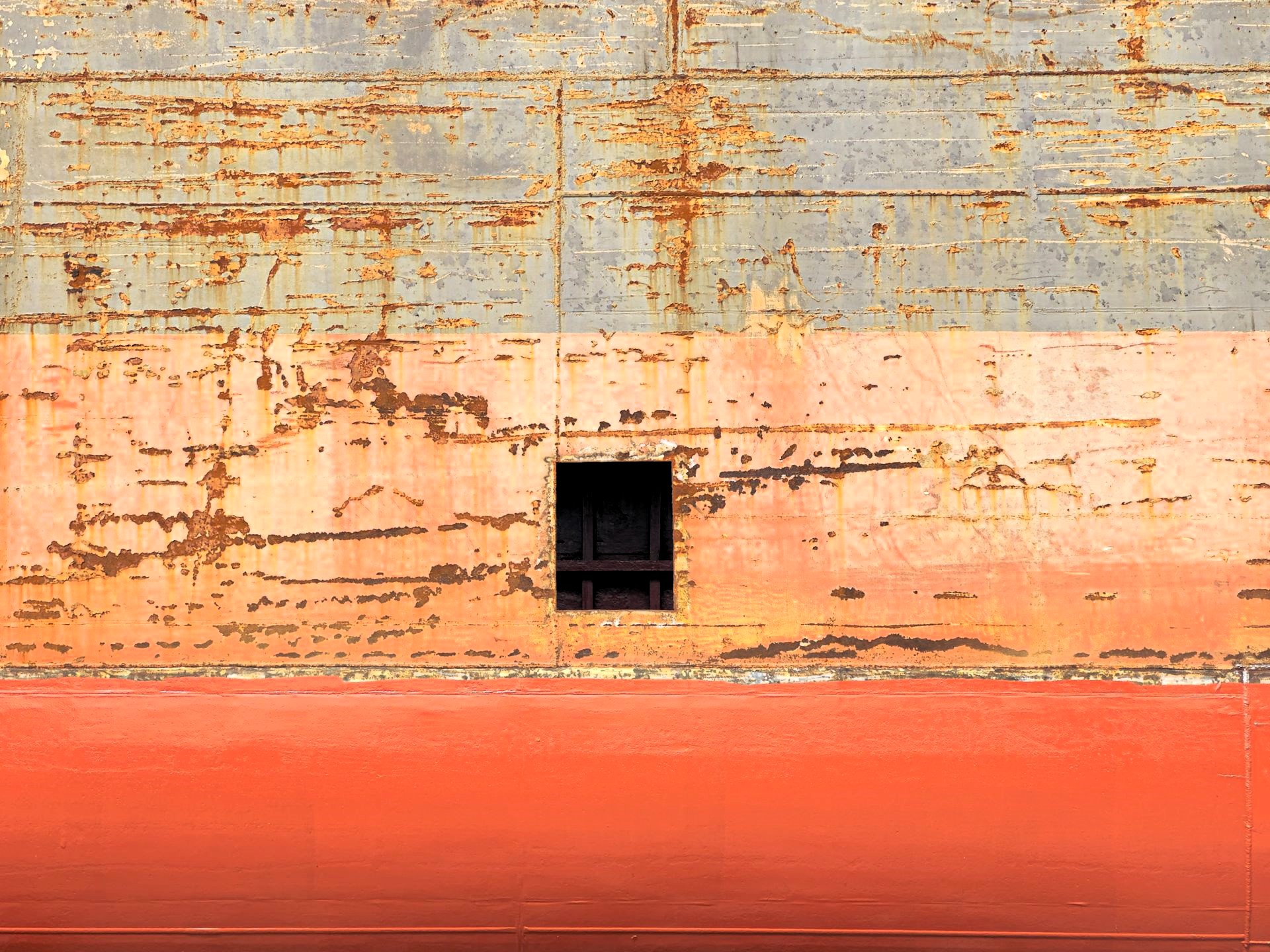 Close-up of a weathered ship’s hull with rust-streaked grey and orange paint and a dark rectangular opening