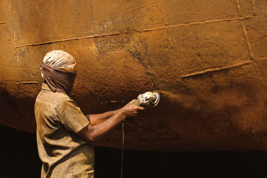 Worker using an angle grinder to remove rust from a ship’s curved steel hull