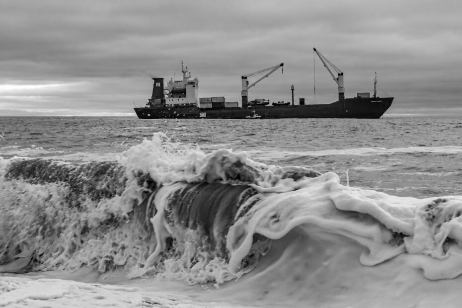 Black-and-white view of a cargo ship with deck cranes beyond a breaking wave beneath an overcast sky