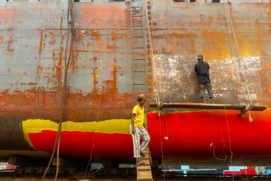 Two workers on wooden platforms beside a ship’s hull, with rust patches, exposed metal, and red and yellow paint