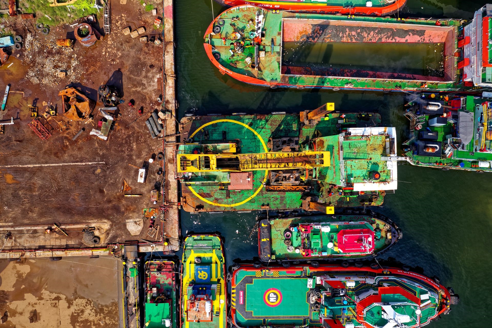 Overhead view of colourful tugboats and work vessels moored beside an industrial quay, with yellow lifting equipment on deck