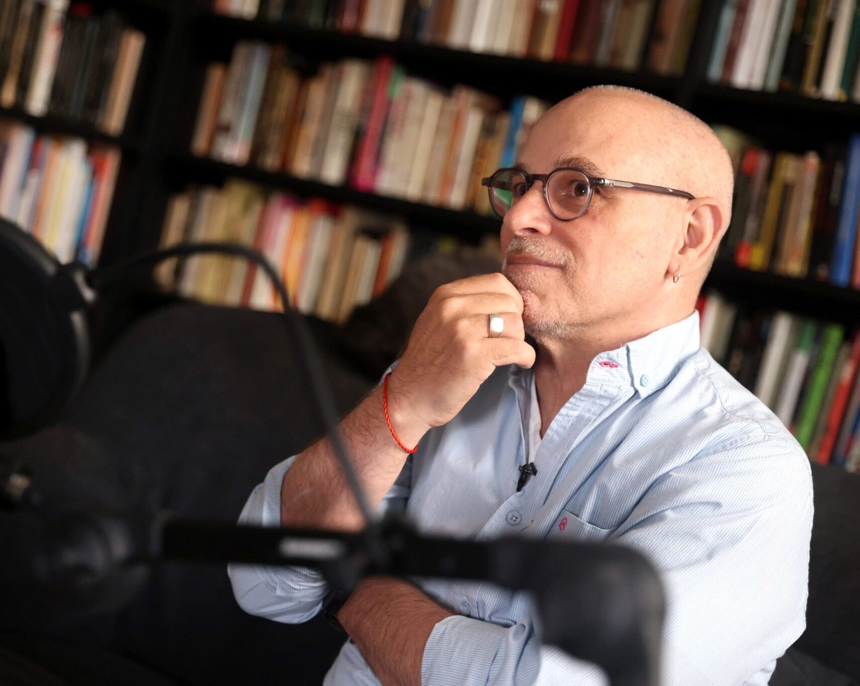 Gadi Taub, wearing round glasses and a light blue shirt, sits thoughtfully before bookshelves, with a podcast microphone in the foreground