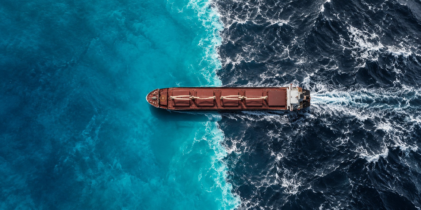 Aerial view of a bulk carrier crossing a sharp boundary between calm turquoise waters and dark, turbulent seas, symbolising widening regional disparities in dry bulk freight markets
