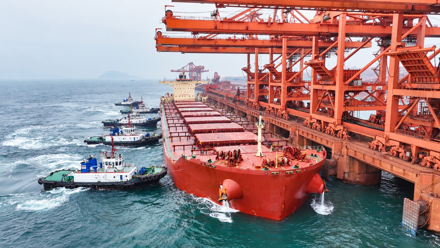 Large red bulk carrier alongside an industrial iron ore terminal, surrounded by tugboats beneath towering orange cranes