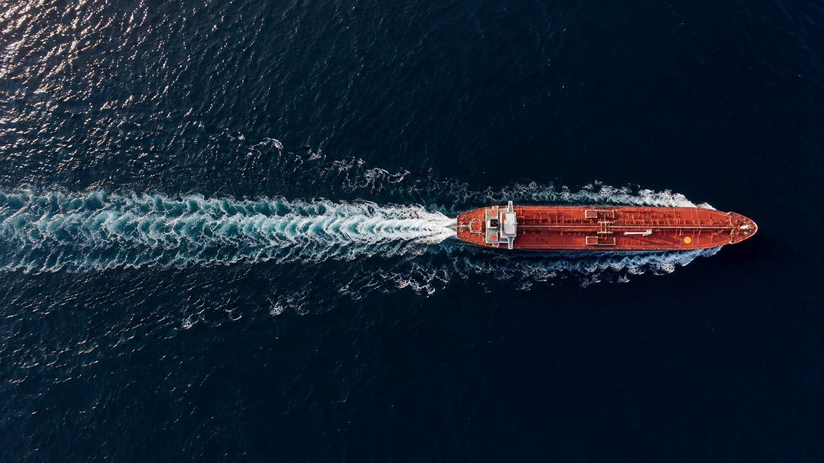 Aerial view of a lone red product tanker crossing dark blue waters, leaving a white wake behind it
